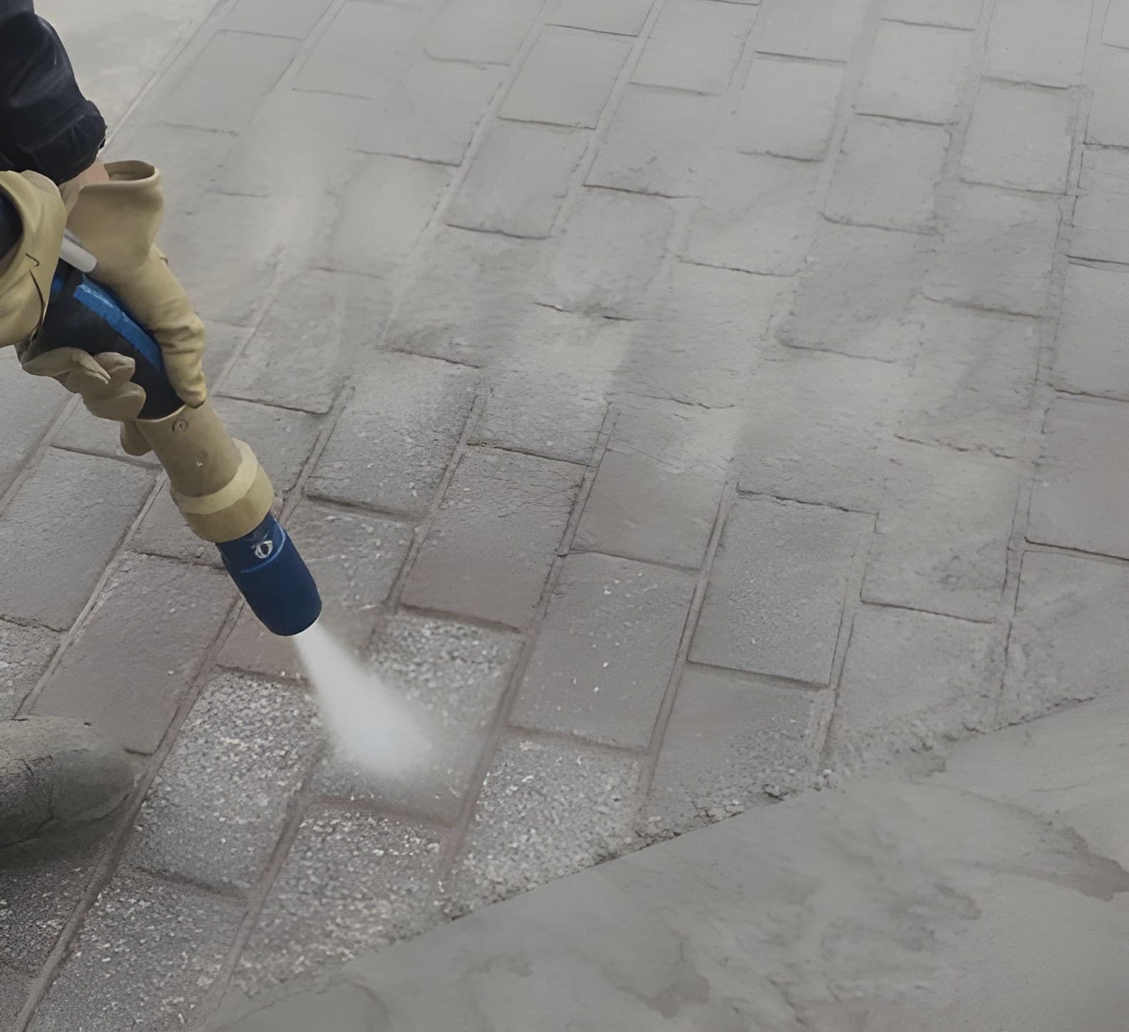 The vapour blasting nozzle mid-pass over pavers — the wet fan of abrasive and the clean line behind it